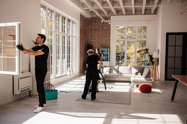 Two people from a professional maid service cleaning a room with a large window, ensuring a tidy and bright space.